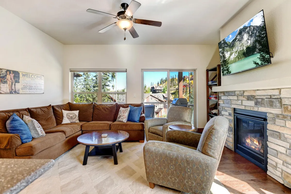 A cozy living room featuring a comfortable sectional sofa centered around a stone fireplace, with large windows offering expansive mountain views. Subtle decor and abundant natural light create a warm, inviting atmosphere, perfect for gathering with family or friends. The HD smart TV is mounted above the fireplace, and plush throws add to the comfort, subtly referencing space for board games or movie nights.