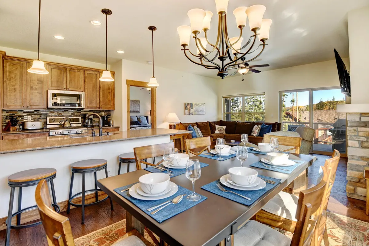 A stylish dining area anchored by a large table set for a meal, with a chandelier overhead and the kitchen visible in the background. The setup is ready for family dinners or festive holiday gatherings, reinforcing the communal aspect of the space.