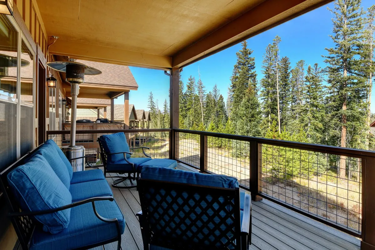 A tranquil wooden deck set amidst green trees, featuring blue lounge chairs and a sturdy patio table. The setting is peaceful and inviting, perfect for enjoying morning coffee or evening wine, with enough space for small gatherings or quiet contemplation. The natural backdrop underscores the Rocky Mountain setting and the connection between interior and exterior living.