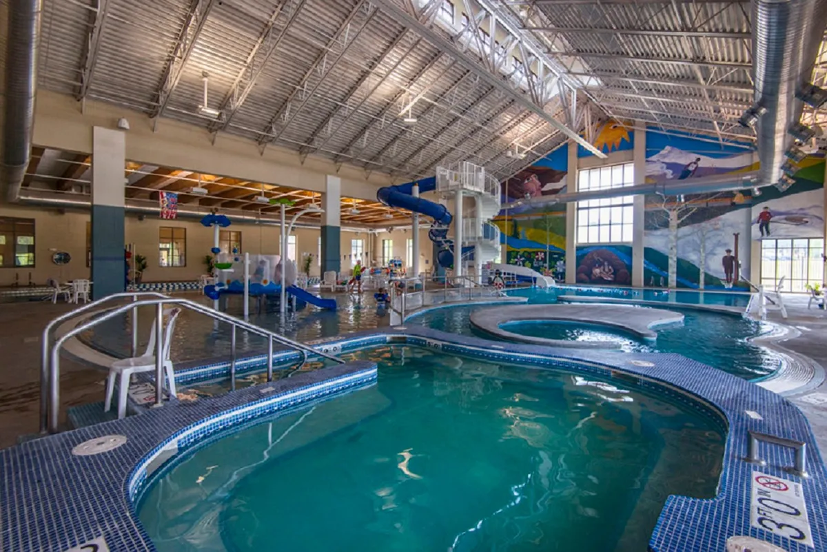 A vibrant indoor recreation center pool area with a swimming pool, water slide, and children playing. The colorful, mural-adorned space exudes energy and fun, suggesting a family-friendly amenity for relaxation and play.