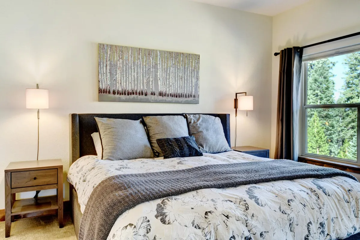 A serene bedroom with a well-made bed, tasteful artwork on the wall, and a window offering a glimpse of nature outside. The space looks peaceful and is filled with soft natural light, emphasizing comfort and relaxation.