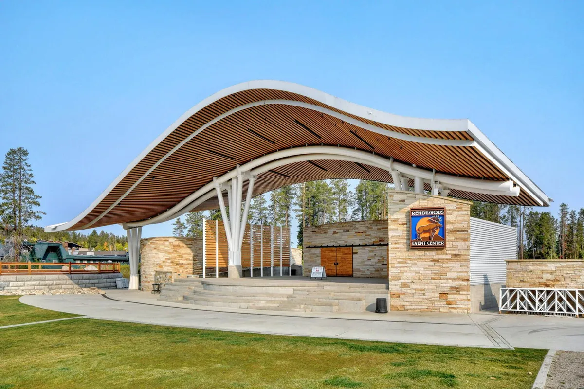 An open-air stage surrounded by stone walls and a curved roof structure, set in a community park. The stage area is designed for concerts and community events, with a casual outdoor seating area that can be filled by guests during performances. Greenery and open sky complete the festive, communal vibe.