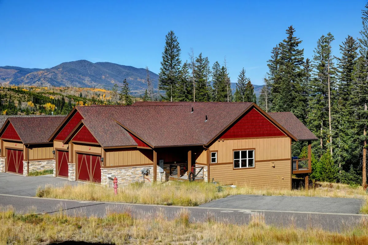 The image showcases a luxurious mountain chalet nestled among tall trees, with a dramatic mountain backdrop and a sweeping view of the Colorado wilderness. The home’s inviting exterior glows in the soft sunset light, suggesting serenity, privacy, and the promise of memorable outdoor gatherings. Wildflowers dot the landscape, while the expansive sky hints at incredible stargazing opportunities.