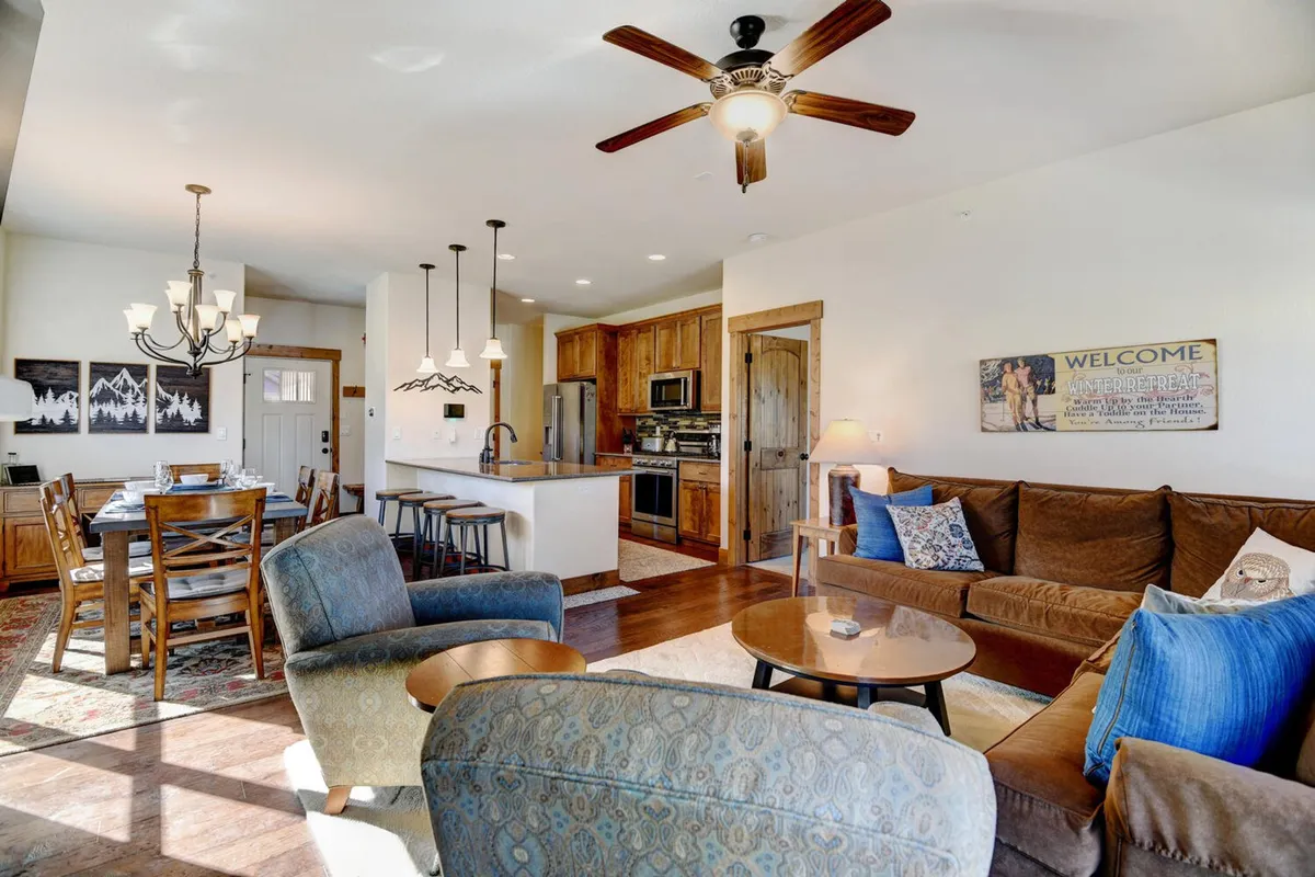 An open-concept space combining a wooden dining table, kitchen island with bar stools, a cozy seating area, and warm lighting. The kitchen is equipped and accessible, while the arrangement encourages both cooking and togetherness.