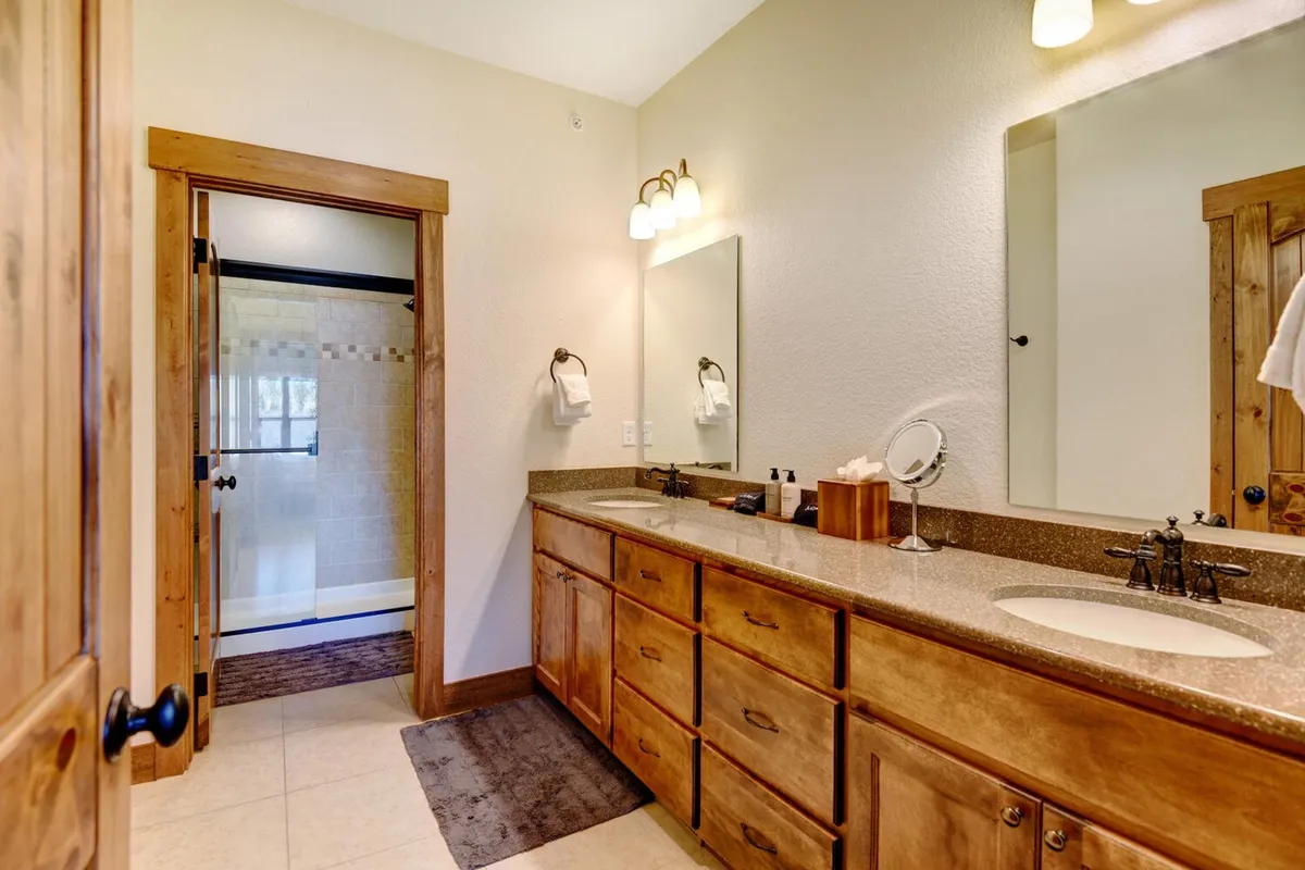 A modern bathroom with a double sink vanity, large mirror, wooden cabinetry, and a glimpse of the shower area. The space is bright, fresh, and exudes a spa-like feel.