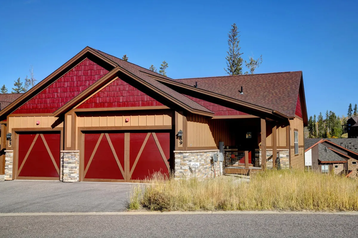 The exterior of a cozy mountain home featuring red garage doors, a textured stone foundation, and wooden accents. The inviting porch and yard suggest spaces for relaxing, playing games, or gathering around a fire pit, all set against a backdrop ideal for both snowy winters and sunny summers.